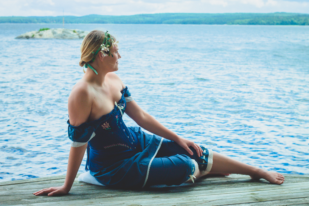 Kvinna i blå korsett på brygga vid vatten. Dräkt av Mytomsydd, Julia Elstring Högberg. Fotograf för Mytomsydd: Johanna Ene